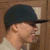 Pixelated close-up of a person wearing a dark blue cap and light-colored shirt against brown background.