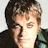 A pixelated close-up of a person with short brown hair, wearing dark clothing against a light background.