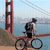 Pixelated image of a person riding a bicycle near a large red bridge structure with water and sky in the background.