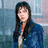 Pixelated image of a person with long dark hair wearing a blue jacket over an orange and white striped shirt against a blurred background.