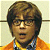 Pixelated portrait of a person with short brown hair, wearing glasses and an orange jacket over a blue shirt.