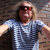 Pixelated static image of a person wearing a light blue striped shirt and dark sunglasses, arms extended outward against a blurred background.