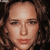 Pixelated close-up of a person's face with light skin tone, dark brown eyes, and reddish lips, set against a dark background with curly hair visible on the sides.