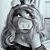 Black-and-white pixelated image of a pig-like puppet with long wavy hair, wearing a dark dress with light patterns and a pearl necklace, positioned against a light background with partial architectural details.