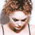 Pixelated close-up of a person with curly brown hair, dark eyes, and a neutral expression, wearing a light-colored top with thin straps.