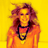 A person with long wavy hair wearing an orange shirt and layered necklaces against a yellow background.