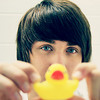 A person with dark hair holds a yellow rubber duck with a red beak near their face against a light background.