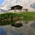 Pixelated landscape with a house on a hill, green grass, and water reflecting the scene under a cloudy sky.