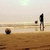 Pixelated beach scene with two figures standing on sand under a pale sky, one figure holding an object near the horizon.