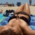A person in a black bikini top lies on a blue patterned towel at the beach, with sand and distant figures visible behind them.