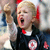 Static pixelated image of a person with blonde hair wearing dark clothing and red accents, mouth open in an expressive pose against a blurred background.