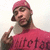 Pixelated image of a person wearing a red beanie and red shirt with dark text, making a hand gesture with the index finger raised.