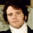 Static pixelated image of a person with dark curly hair, light skin, and a neutral expression wearing a white shirt and dark jacket against a blurred background.
