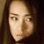 Pixelated close-up of a person's face with long dark hair, neutral expression, and light skin tone against a dark background.