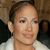 Pixelated close-up of a person with light skin tone, dark hair pulled back, wearing a white top with dark shoulder accents, neutral expression, and visible ear.