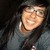 A pixelated close-up of a person with long dark hair wearing glasses and a gray top against a dark background.
