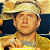 Pixelated image of a person wearing a light-colored cap and yellow jacket, seated with dark blue background elements.