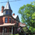 Pixelated image of a multi-story house with a steep roof and chimney, set against a light blue sky with green foliage on the right.