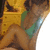 A person in a green top and brown pants sits on a yellow patterned surface with legs crossed, head turned slightly right.