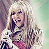 A person with long blonde hair and bangs holds a silver microphone, wearing a light-colored jacket over a red patterned top against a blurred background.