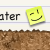 Yellow sticky note icon with winking eye and curved smile, positioned next to partial text on a light background above brown textured ground.
