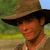 A static pixelated image shows a person wearing a wide-brimmed brown hat with black band, light-colored shirt, and dark necklace against blurred green and blue background.
