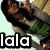 A person with long dark hair wearing a gray cardigan over a green patterned top, holding an object near their face against a light background.