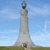 Pixelated static image of a tall stone monument with a spherical top, set against a blue sky with light clouds and green grass at the base.