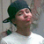 Pixelated image of a person wearing dark green headwear and light-colored clothing, with visible facial features including eyes, nose, mouth, and skin tone variations.