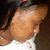 A pixelated close-up of a person's head and shoulder with light brown hair, wearing a white top against a dark background.