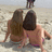 Two figures in swimwear sit on a sandy beach with a third figure standing behind them, all viewed from behind.