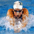 A swimmer in a white cap and goggles performs the breaststroke with arms extended forward, surrounded by blue water and white splashes.