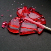 Red translucent heart-shaped lollipop with a wooden stick on a dark gray background, scattered pink fragments around it.