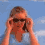 A person in a light blue shirt adjusts dark sunglasses against a blue sky with white clouds, then lowers hands to reveal a neutral expression.