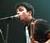 Pixelated image of a person singing into a microphone with dark hair and light skin, wearing a sleeveless top against a blurred background.
