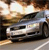 A silver sedan with dark tinted windows is positioned on a road, viewed from a low angle with a blurred background of warm-toned light and dark shapes.