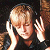 Pixelated image of a person with light brown hair holding a phone to their ear and raising a hand in a gesture, set against a dark background.