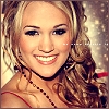 A static image of a smiling person with long wavy blonde hair, wearing a red strap and a dark beaded necklace against a blurred background with light speckles.