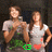 Two women in white shirts pose against a dark background with green foliage; one woman holds an object while the other has hands on hips.