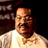 Pixelated static image of a person in a light-colored shirt against dark background with visible dithering and compression artifacts.