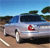 Pixelated image of a light-colored vehicle with a red emblem on the side, positioned on a dirt surface under a blue sky with green foliage in the background.