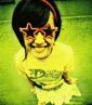 A person with short dark hair wears star-shaped sunglasses and a yellow shirt with green text, standing against a blurred green background.