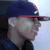 Pixelated profile view of a person wearing dark blue cap with red brim and small white emblem, light gray skin tone, black clothing against pale blue background.