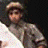 Pixelated figure in light-colored flat cap and white shirt against dark background, cropped to upper body with visible shoulder and arm.