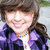 A person with brown hair and a purple jacket smiles in a static image with a blurred outdoor background.
