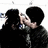 Two figures with black hair embrace closely, one figure's hand touches the other's face in a gentle gesture.