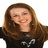 A pixelated static image of a person with long brown hair wearing a black top with white accents, smiling with eyes partially closed against a white background.