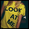 A person wearing a yellow short-sleeve shirt with black text reading LOOK A7 ME, with visible arms and hair against a dark background.