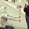 A person in a light gray hoodie kneels on one knee holding a bouquet of red flowers toward another person standing on a paved street.