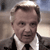 Static pixelated image of a person in a light-colored suit jacket, white shirt, and dark tie, with a blurred background showing indistinct shapes and muted tones.